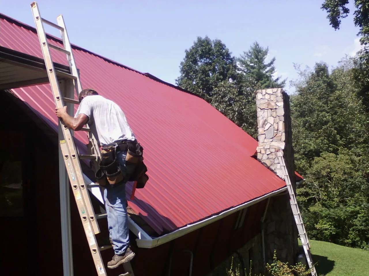 Roofer on a ladder beside a red metal roof and stone chimney — Soffit & Fascia Repair by Roofing Brothers in Coopersville